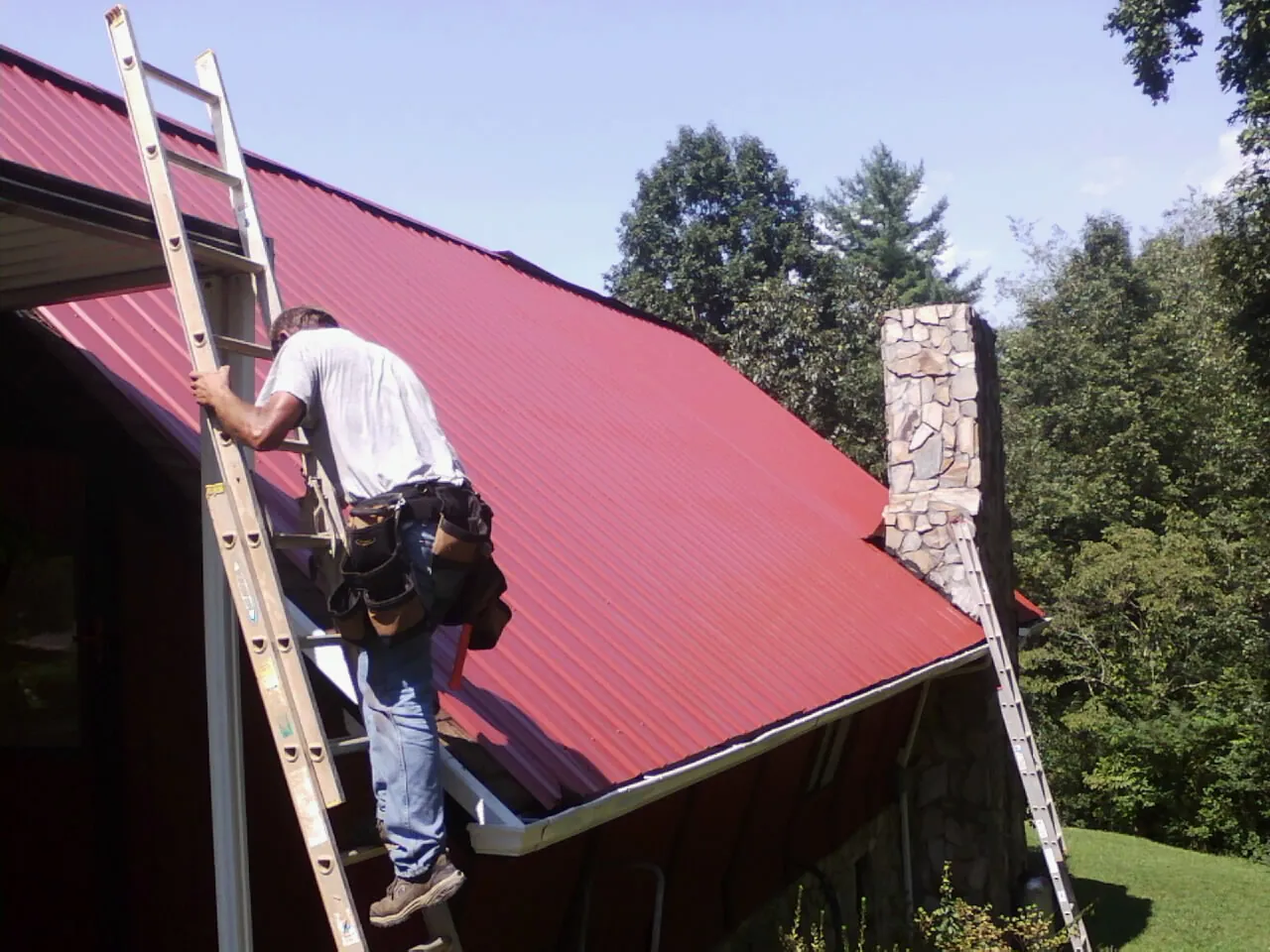 Roofer on a ladder beside a red metal roof and stone chimney — Soffit & Fascia Repair by Roofing Brothers in Enderlin